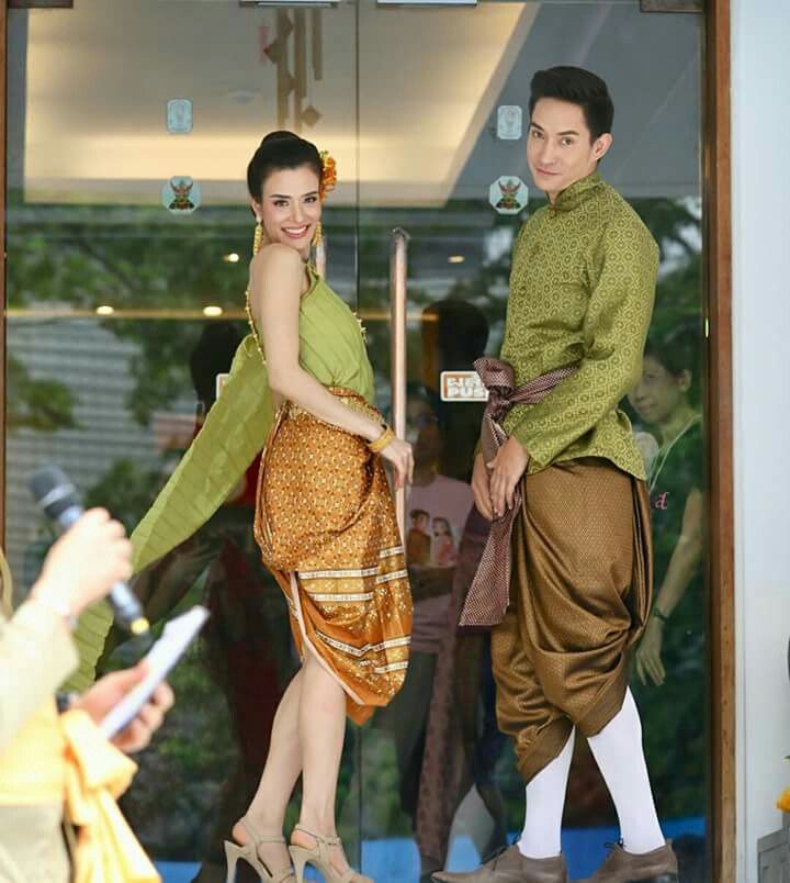 a thai couple dressed in traditional clothing stand together outside a building, smiling.  the man is wearing a green patterned shirt and brown pants, and the woman is wearing a green dress and a brown patterned skirt.
