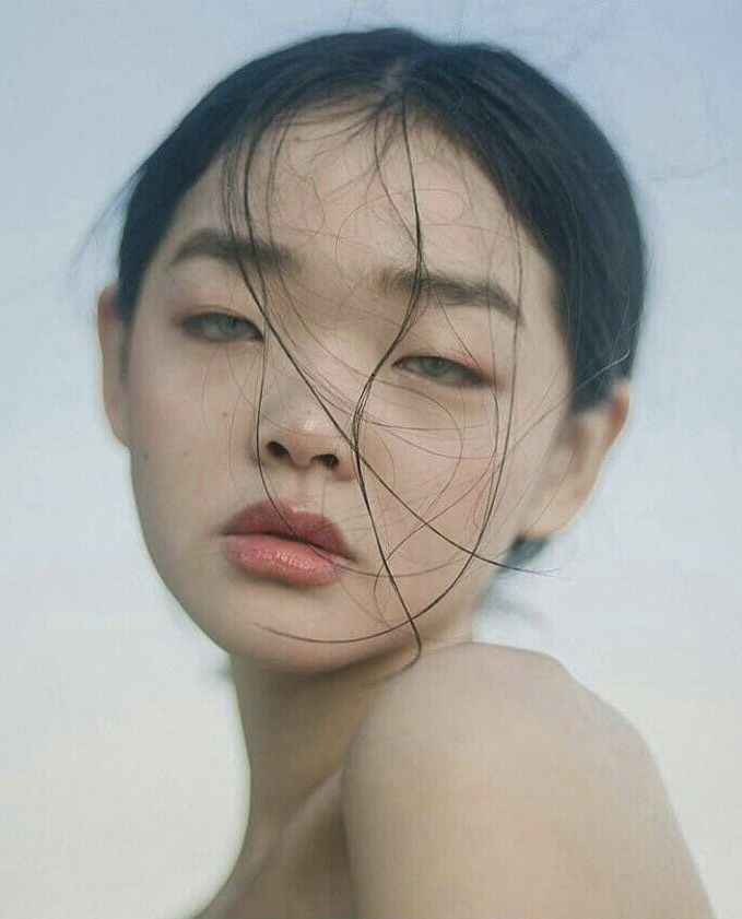 a closeup portrait of a young woman with wet, dark hair partially obscuring her face. she has pale skin, a neutral expression, and minimalistic makeup. loose strands of fine hair are scattered across her face. the image focuses on her face and shoulders.