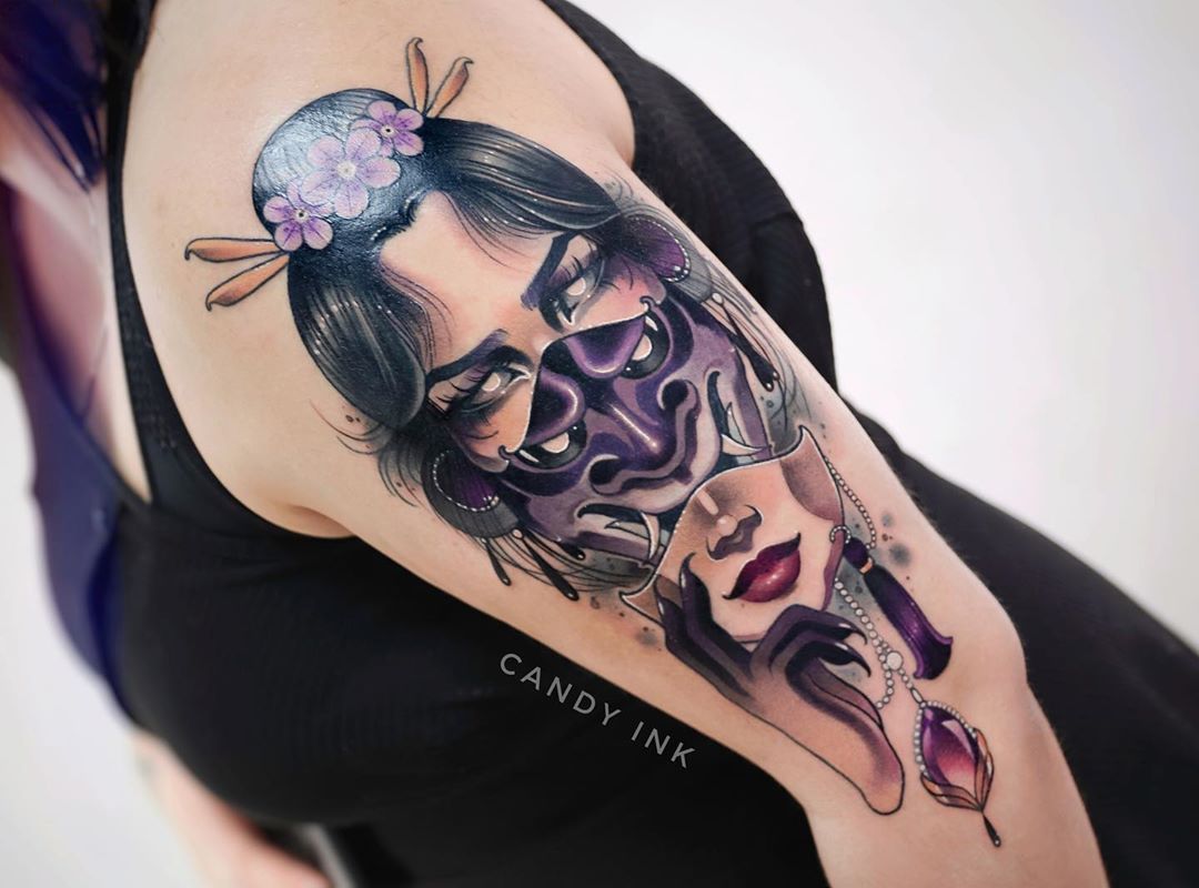 a closeup photograph showcasing a vibrant tattoo on a person's upper arm. the tattoo, rendered in a realistic style with bold lines and shading, depicts a woman with dark hair and purple flowers in her hair, holding a fierce oni mask in front of her face. the mask is rendered in shades of purple, black, and grey, with sharp teeth and horns. the woman's visible eye stares directly at the viewer, creating a sense of mystery and intrigue. the tattoo is detailed and wellexecuted, demonstrating a high level of skill from the artist. the soft lighting and closeup perspective highlight the tattoo's intricate details and vibrant colors.