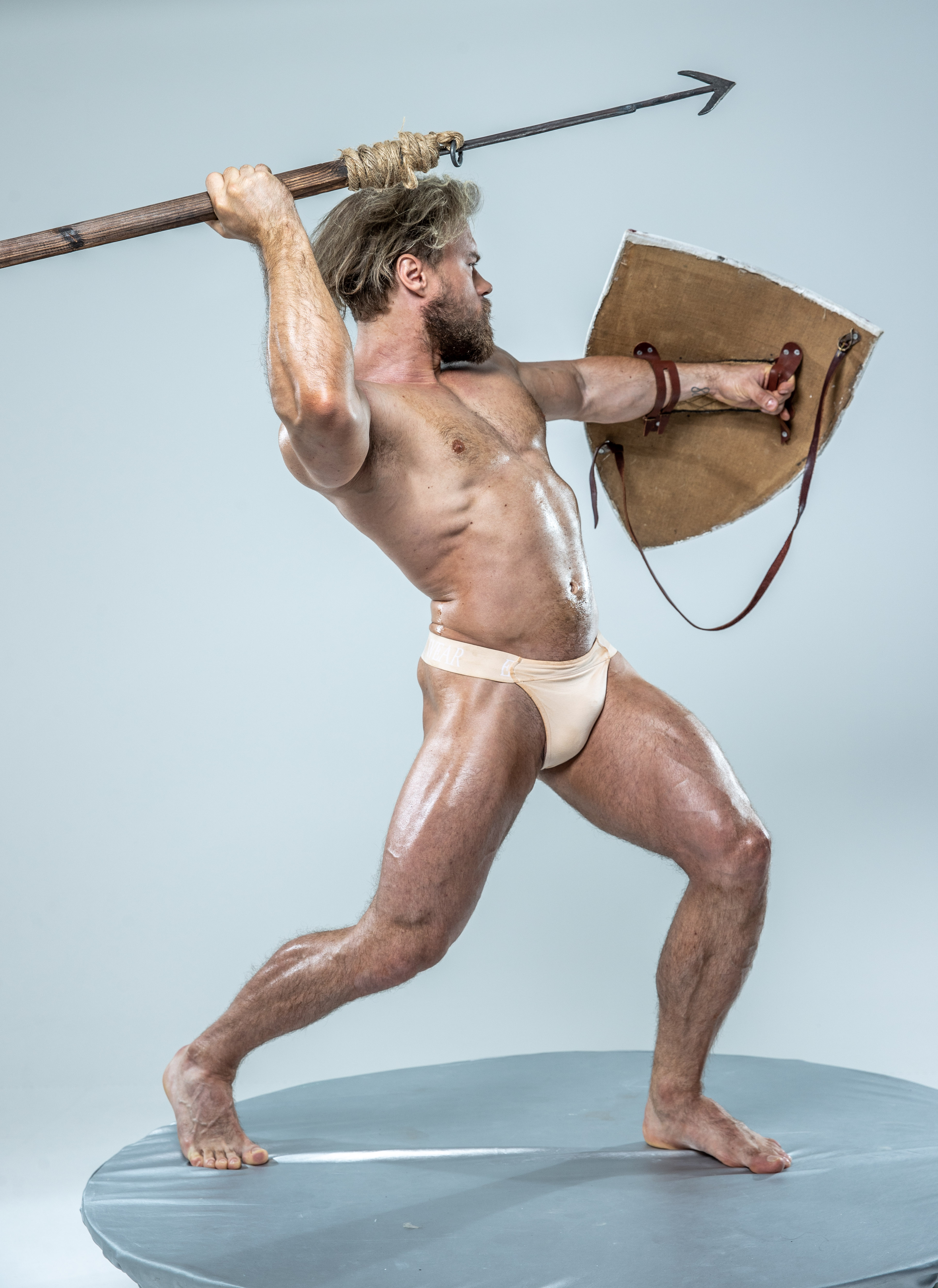 a muscular man with long blond hair and beard, wearing only a loincloth, stands in an attack pose against a light gray background. he holds a spear aloft in his right hand, ready to strike, and a round shield in his left hand for defense. he is illuminated with bright studio lighting, highlighting his physique and the texture of his skin. the scene appears staged, suggesting a historical warrior or gladiator.