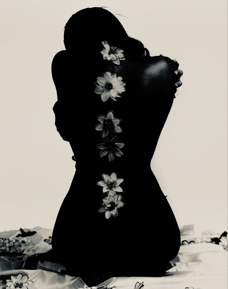 the image is a black and white, almost entirely silhouette style photograph of a woman sitting.  her back is to the camera, and a line of flowers is depicted along her spine.  the lighting is dramatic, focusing on the stark contrast between the dark figure and the lighter background. the overall mood is artistic and evocative, with a slightly sensual and mysterious tone. the woman's pose is somewhat introspective and closed off. the flowers add a touch of delicate detail against the strong silhouette. the bottom of the image shows a hint of fabric or textile beneath the woman.