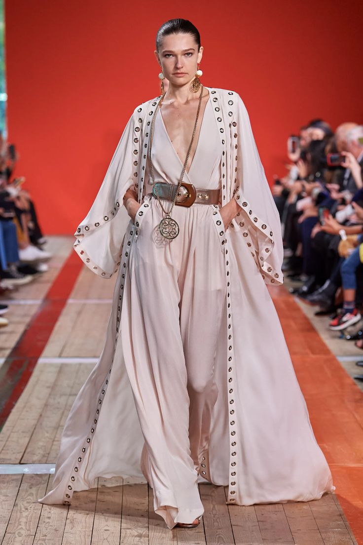 a female model walks the runway in a chic, long, white dress. the dress has long sleeves, a deep vneck, a belt, and a flowing skirt. the model is also wearing  jewelry,  and sandals. the model is walking with confidence, showcasing the designer's elegant and stylish collection.
