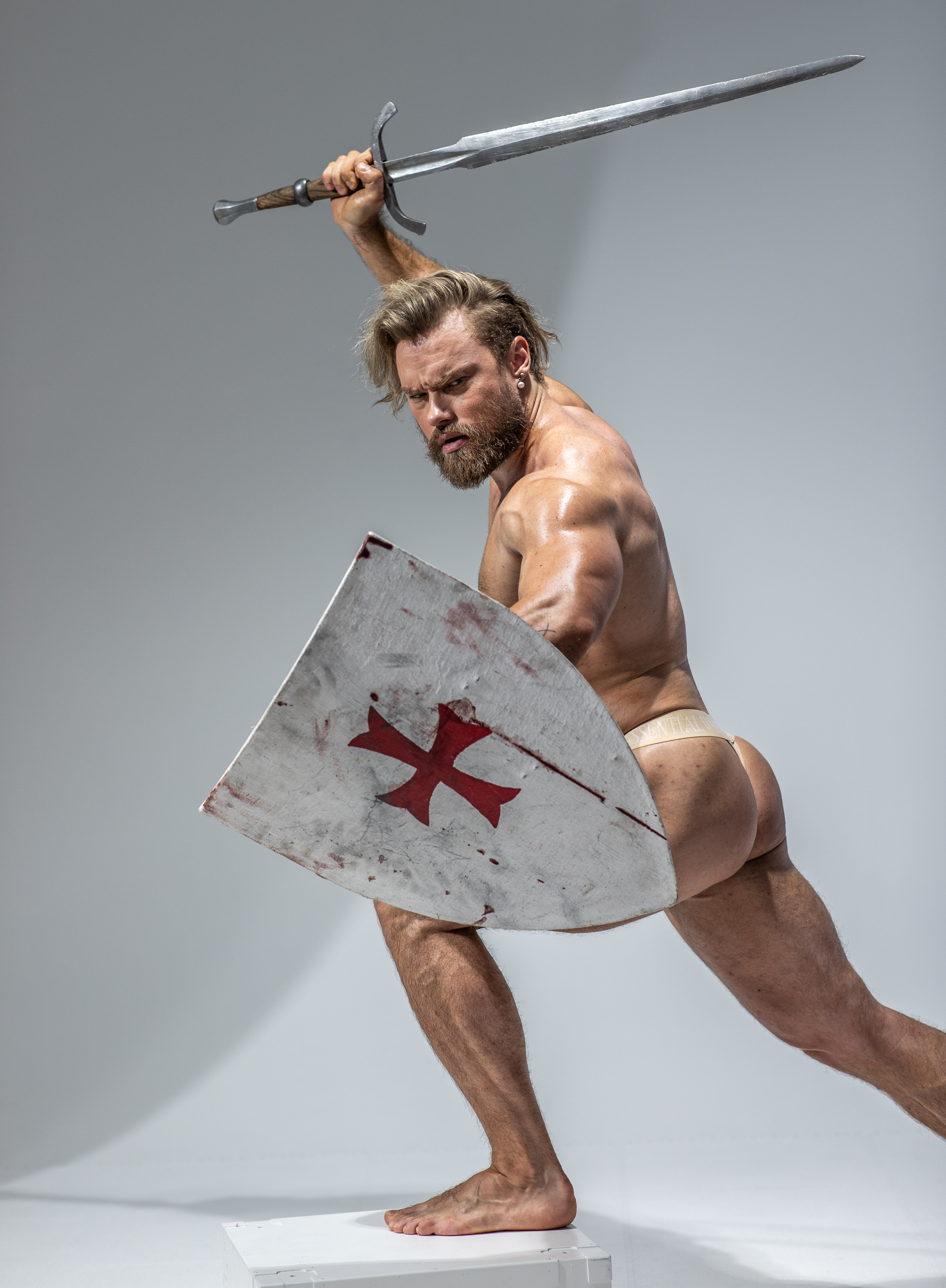 a muscular man with long blond hair and a beard poses dynamically against a light gray background. he's depicted as a warrior, perhaps a knight or crusader, wielding a large sword in his right hand raised above his head and a shield in his left. he's barefoot and wears only a beige loincloth. the scene suggests action and combat, frozen in a dramatic moment.