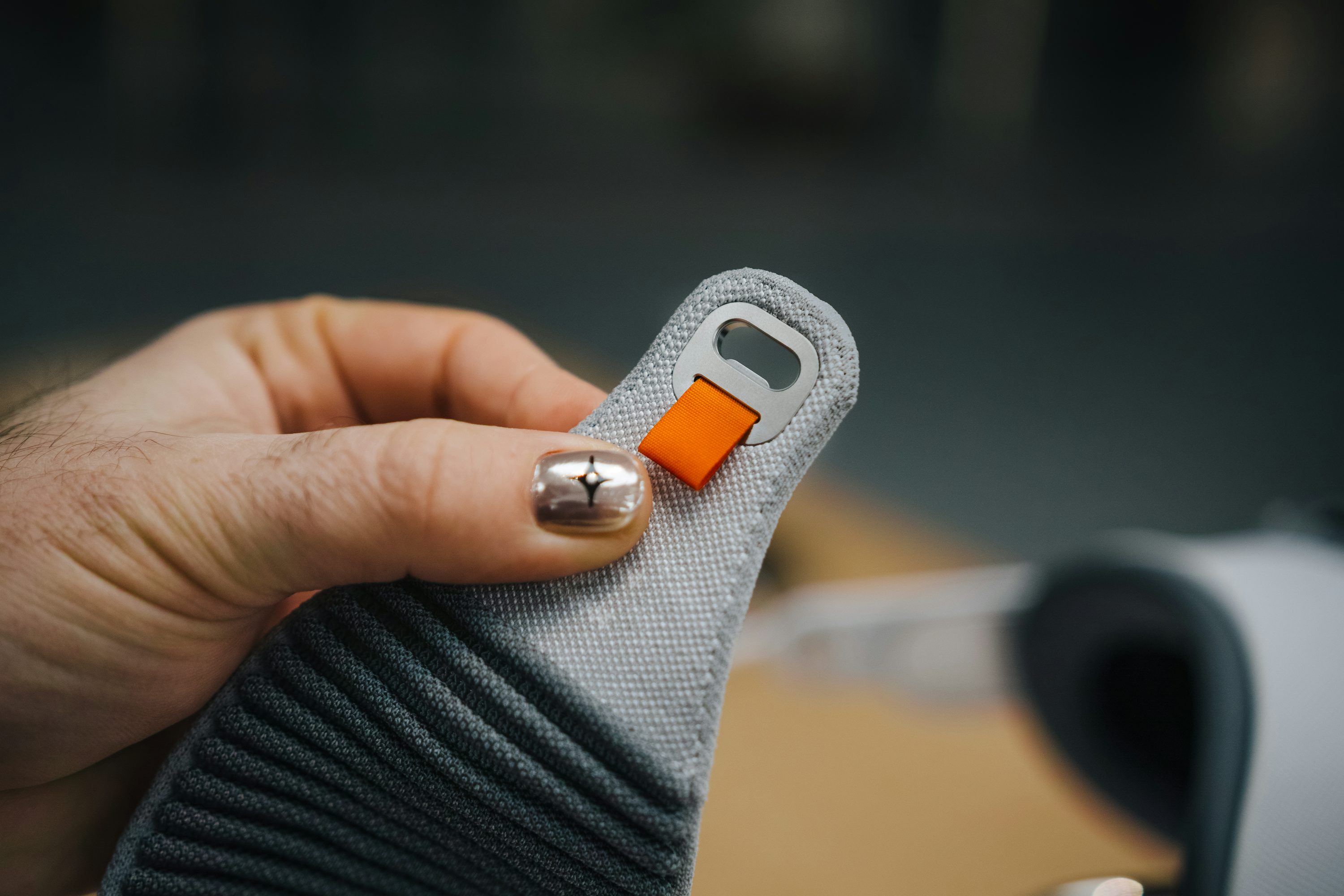 a hand with a painted fingernail holding a gray fabric strap with a metal ring and an orange fabric tag. the strap has a ribbed pattern on one side. a blurry background shows another object, potentially a pair of glasses.