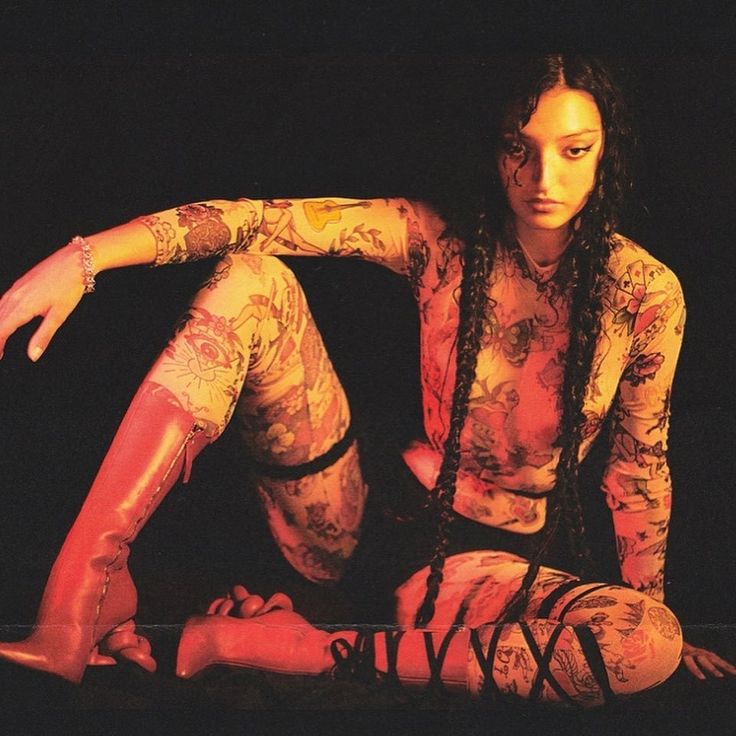 a woman with long, dark, braided hair sits on a dark background wearing a sheer, longsleeved shirt and leggings that have a tattoo design all over them. she is wearing red boots and has a large, dark tattoo on her thigh. she has dark eye makeup. the image appears to have been shot in a studio with a single, strong light source. the background is dark and mostly out of focus.