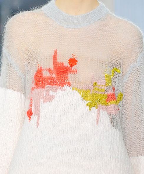 a close up of a model wearing a white, looseknit sweater with colorful abstract embroidered designs on the front. the sweater has long sleeves and a crew neck. the model is standing with the sweater draped on them.  the image is taken from the front.  the background is blurred and out of focus.