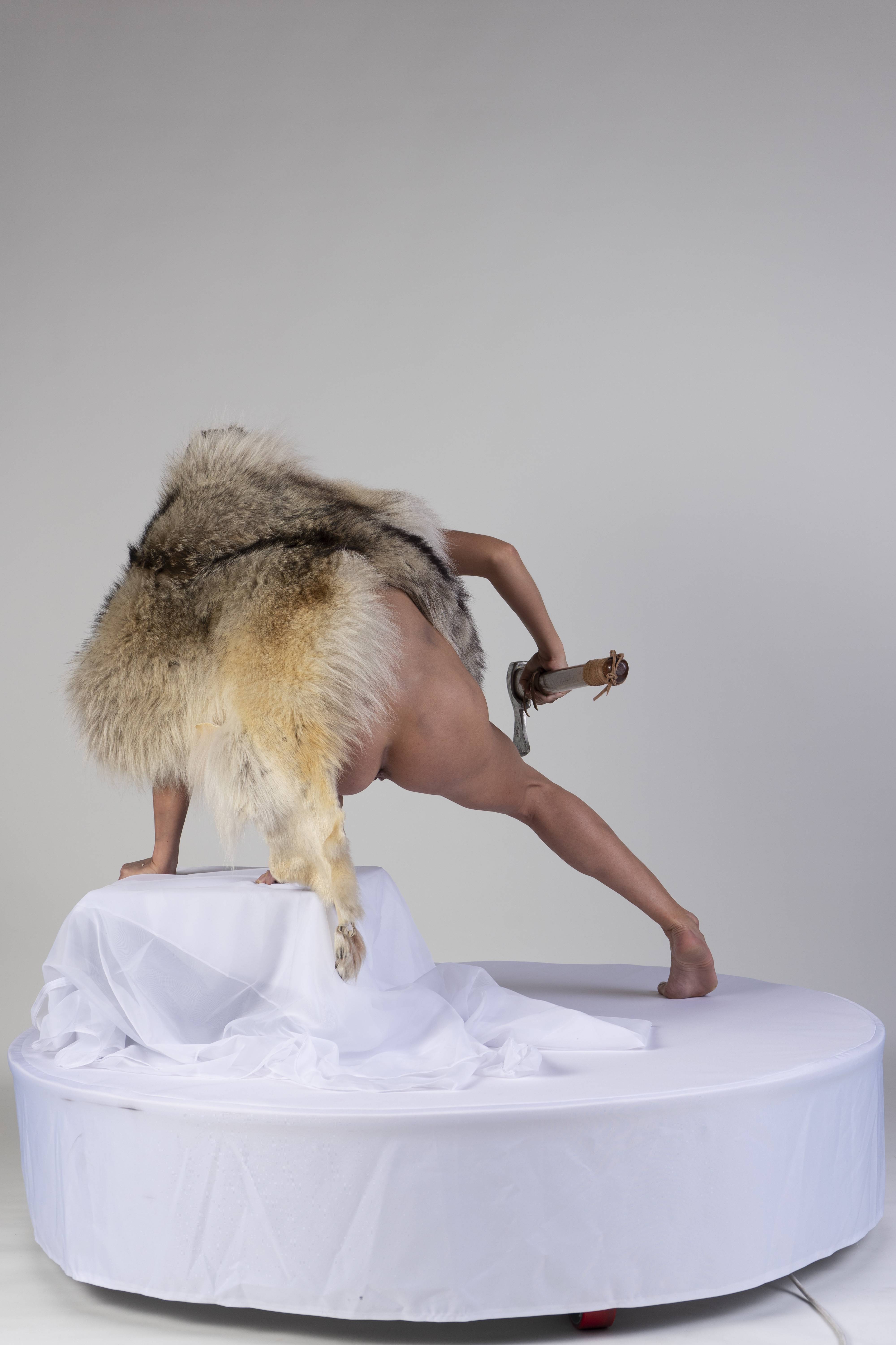 a nude person draped in a large animal fur is standing on a white platform in front of a white backdrop. the person is in an unconventional pose, with one hand and foot on a smaller platform covered in white fabric, and the other foot on the larger platform. they are holding an axe in their hand. the image is provocative and could be considered avantgarde performance art.
