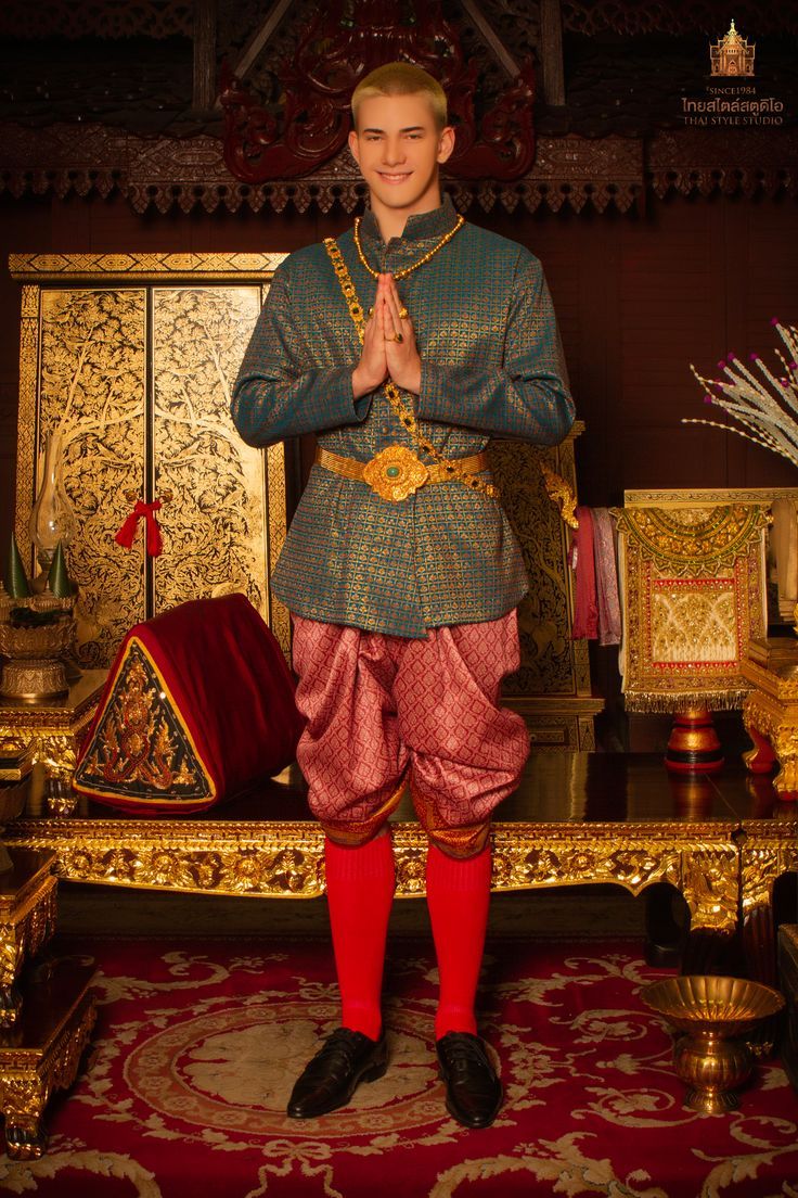 a man in traditional thai clothing stands with his hands clasped together in a gesture of greeting in front of an ornately decorated throne and table. he wears a green and gold brocade shirt,  pink  pants,  red socks, and black shoes. he stands in a traditional pose and looks directly at the camera.  the background of the image features an ornate throne with gold accents and a red and gold rug. the man is dressed in traditional thai clothing, and he is smiling. the image is likely taken in a thai palace or temple.