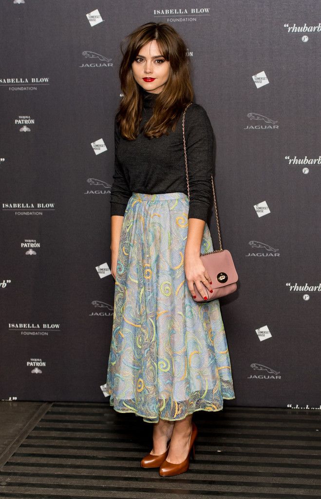 jenna coleman is captured in a fulllength shot, attending an event. she is elegantly dressed in a dark grey turtleneck top paired with a flowing, patterned midi skirt in teal, blue, and cream hues.  her brown leather heels and a dusty rosecolored handbag complete her look. her dark brown hair is styled with bangs framing her face. the background showcases logos and text, including "isabella blow foundation" and "jaguar", suggesting the event's sponsors and possibly its name.