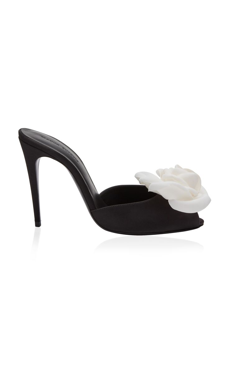 a closeup shot of a black satin heeled mule with a white flower on the top. the shoe has a stiletto heel and an open toe. the shoe is sitting on a white background.