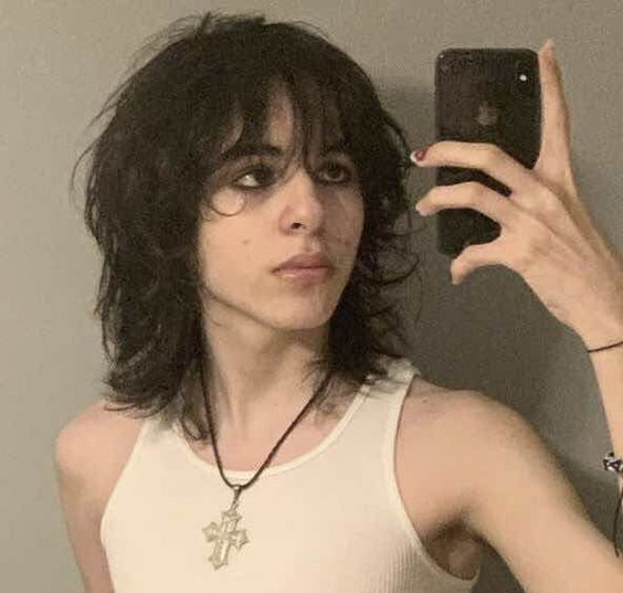 a young person is taking a selfie in a mirror. they have long, dark wavy hair with bangs that are parted in the middle. they are wearing a simple white tank top and a cross necklace. they are looking at the camera with a neutral expression.