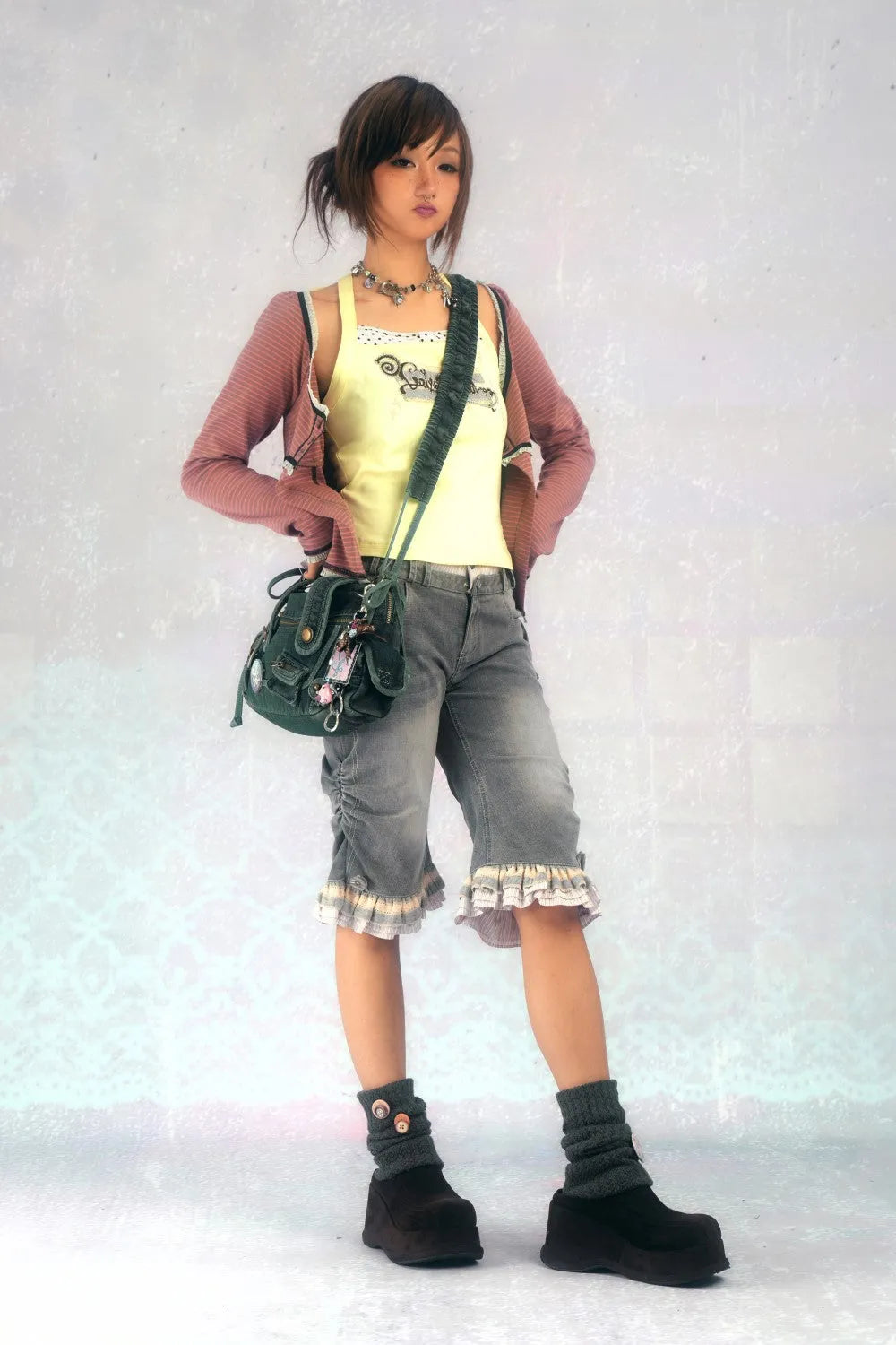 a fullbody shot of a young asian girl posing against a light grey wall. she's wearing a pastel pink cardigan over a yellow tank top, grey denim shorts with ruffles at the hem, dark ankle boots, and dark grey kneehigh socks. she has a shoulder bag slung across her body and is wearing a necklace. her hair is styled in a side ponytail with bangs. the overall style is casual and slightly vintageinspired.