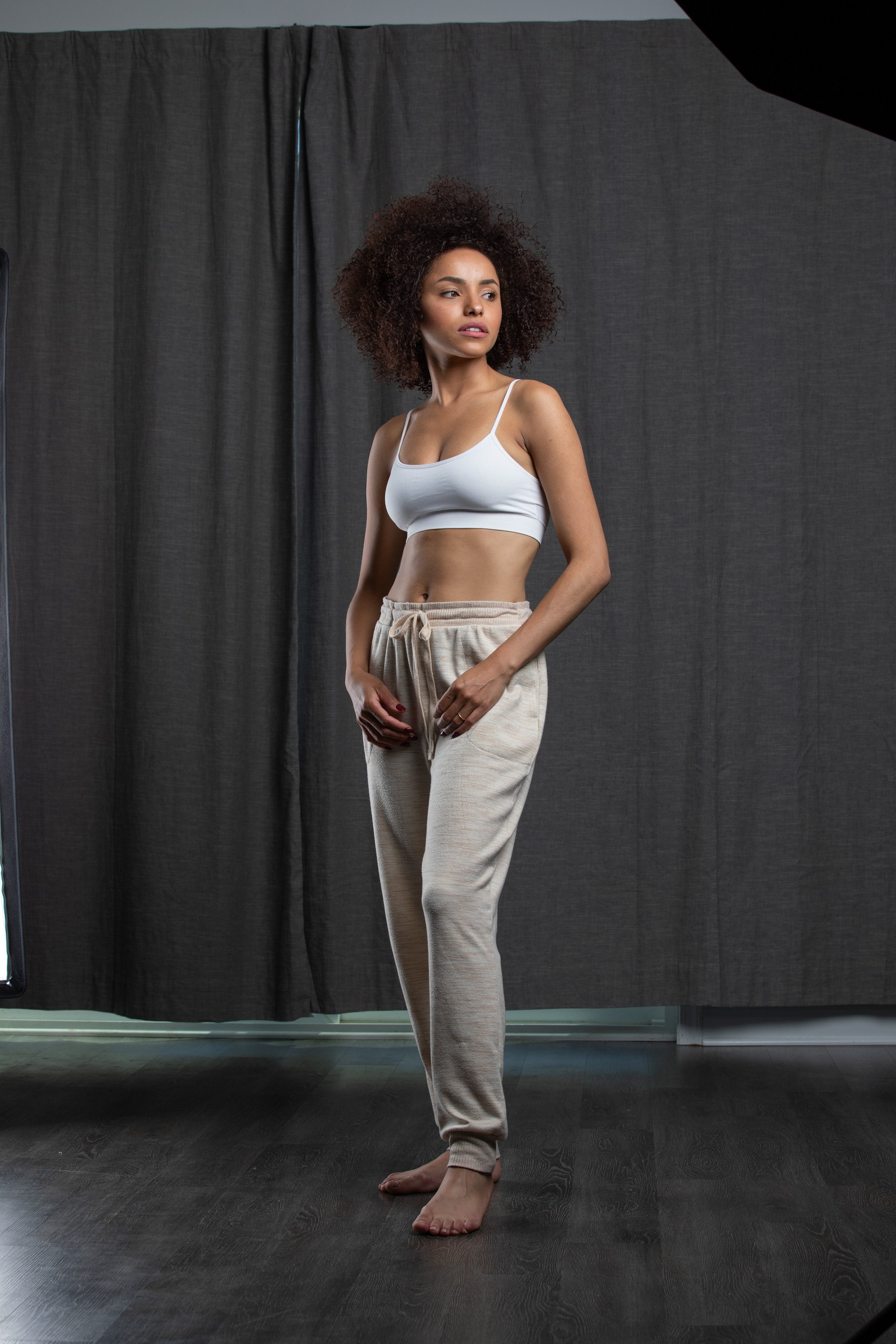 a woman with an afro hairstyle is standing in a studio setting. she is wearing a white sports bra and beige sweatpants. the background is a dark gray curtain. the lighting is soft and diffused, creating a flattering portrait.