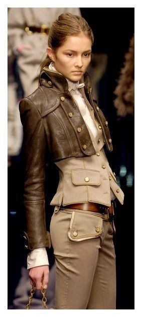 a woman is posing on a runway in a unique fashion outfit. she is wearing a short, brown leather jacket with gold buttons. underneath the jacket is a beige vest, also with gold buttons and pocket detailing. she is also wearing beige pants with pocket details and a brown belt. she is holding a gold chain in her hand.