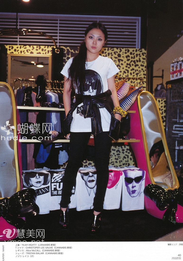 a young woman with long black hair is standing in a clothing store. she is wearing a white tshirt, black pants, and black shoes. the tshirt has a picture of a woman's face on it. the store is decorated with leopard print and pink. there are clothes racks behind her and a mirror on the left side of the image. the image appears to be from a magazine.