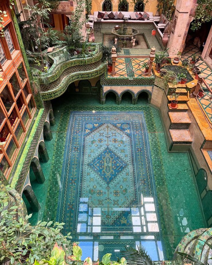 overhead shot of a beautiful, luxurious courtyard. in the center of the courtyard is a pool, decorated with a beautiful, large ruglike pattern on the floor. the pool is surrounded by decorative tile work, mosaics, and plants. a small staircase leads to the pool. around the pool are arched doorways and windows. in the background is a seating area with couches and pillows. the overall atmosphere is serene and luxurious.