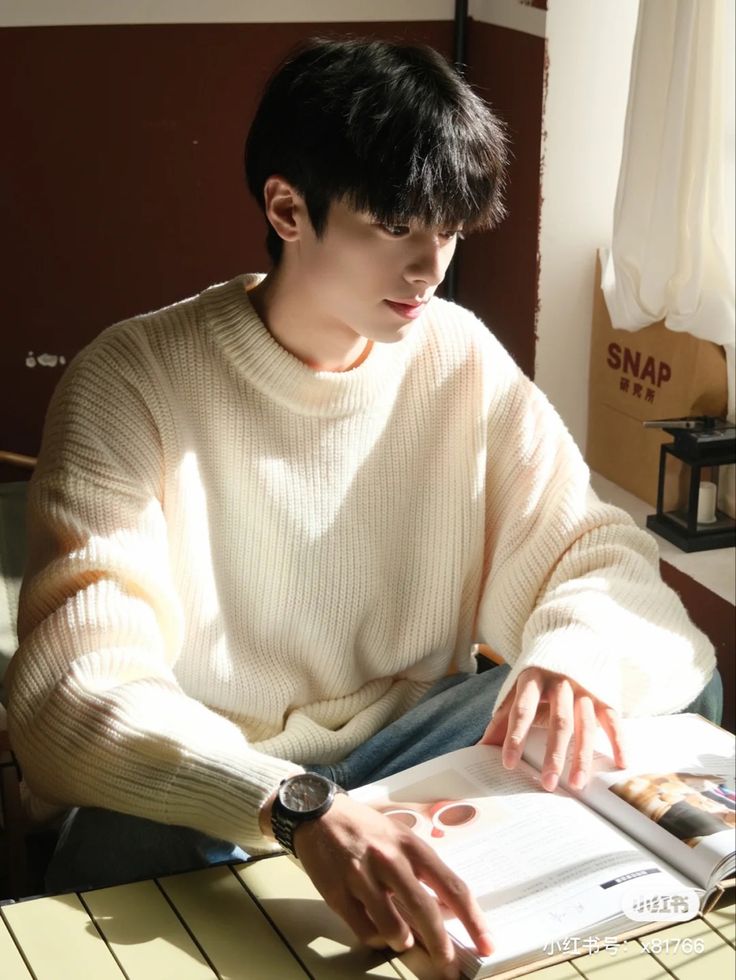 a young man with dark hair and a white sweater sits indoors, illuminated by natural lighting. he is looking down at an open magazine while wearing a wristwatch. the environment appears warm and inviting.