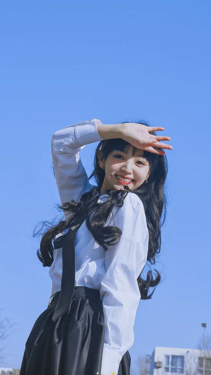 a young woman is smiling, dressed in a school uniform with a black skirt, white shirt and black tie. her hand is raised over her eyes, as if shielding them from the sun. the sky behind her is clear and blue.