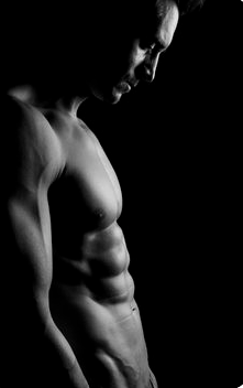 a black and white photograph of a muscular man's torso. the image focuses on the definition of his muscles, highlighted by strong studio lighting that casts deep shadows. the man is standing in profile, his face turned slightly down and out of the frame, creating a sense of contemplation or intensity.