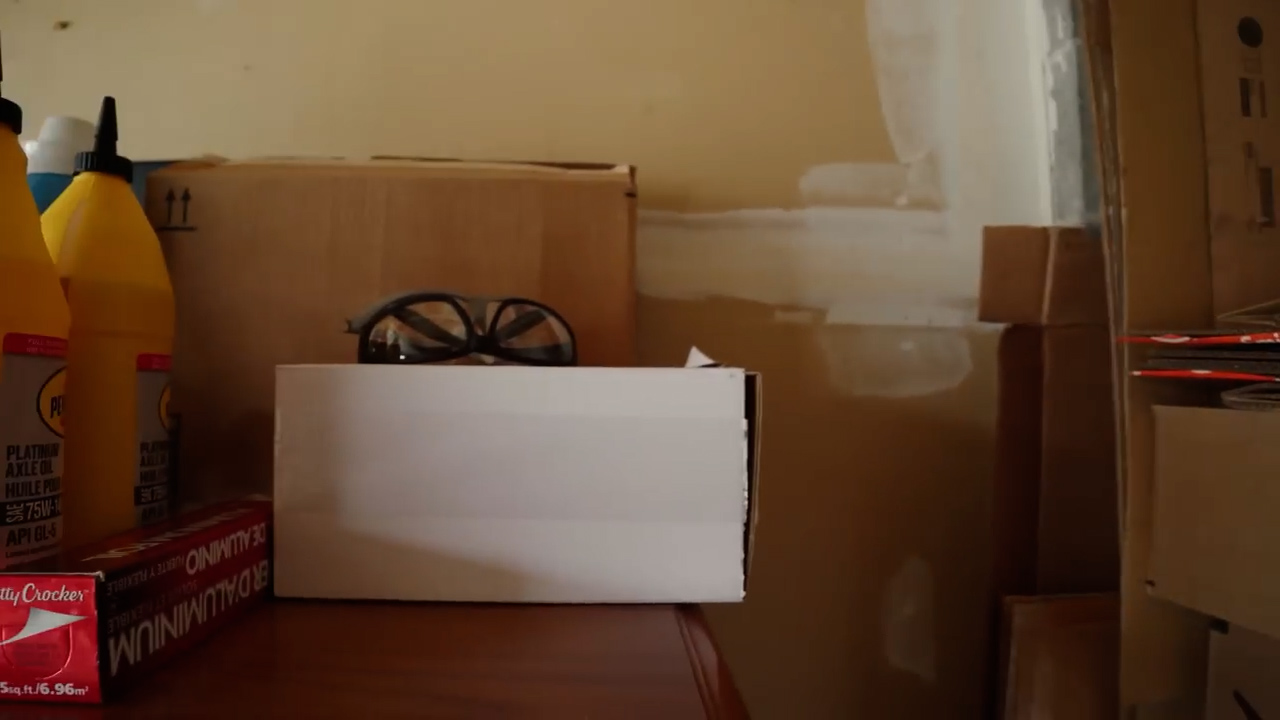 a white cardboard box sits on a table with a pair of safety glasses resting on top.  behind the table are more cardboard boxes and some bottles of liquid. the wall is partially painted white.