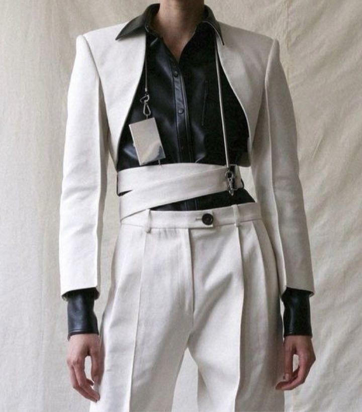 a fashion model is wearing a white suit with a black leather jacket underneath. the jacket is cropped and tied with a white belt that is secured with silver hardware and a chain. the model is posing with their hands on their hips, with their shoulders relaxed and their head slightly tilted to the side.