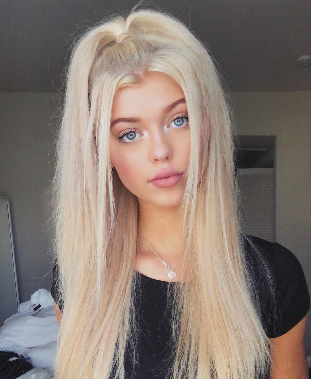 a closeup portrait of a woman with long, blond hair styled in a halfup ponytail. she has striking blue eyes and fair skin. she is wearing a black shirt and a delicate necklace. she is wearing makeup, including lipstick. the background suggests she is in a bedroom, with a bed, mirror, and ceiling light visible.