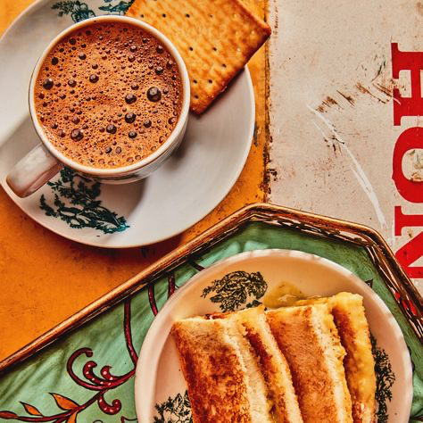 the image is a closeup shot of a classic singaporean or malaysian breakfast: kaya toast and kopi.  a plate of goldenbrown kaya toast (bread spread with kaya coconut jam and butter, then grilled), is presented next to a cup of kopi (coffee).  a crispy cracker rests on the saucer of the kopi cup.  the food is displayed on a vintagestyle tray with a teal and gold patterned surface, against a backdrop of a worn, orange and red wall with faded signage. the overall aesthetic is nostalgic and evocative of a traditional coffee shop setting.