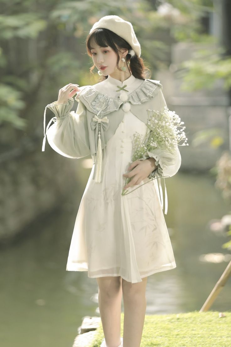 a young woman wearing a delicate, flowing light green and cream dress stands outdoors in a parklike setting. she is posing with a bouquet of baby's breath, and the soft natural light emphasizes the ethereal and feminine feel of the image.  the dress features lace, embroidery, and a layered design with puffy sleeves, indicative of lolita fashion or modern interpretations of traditional chinese clothing.