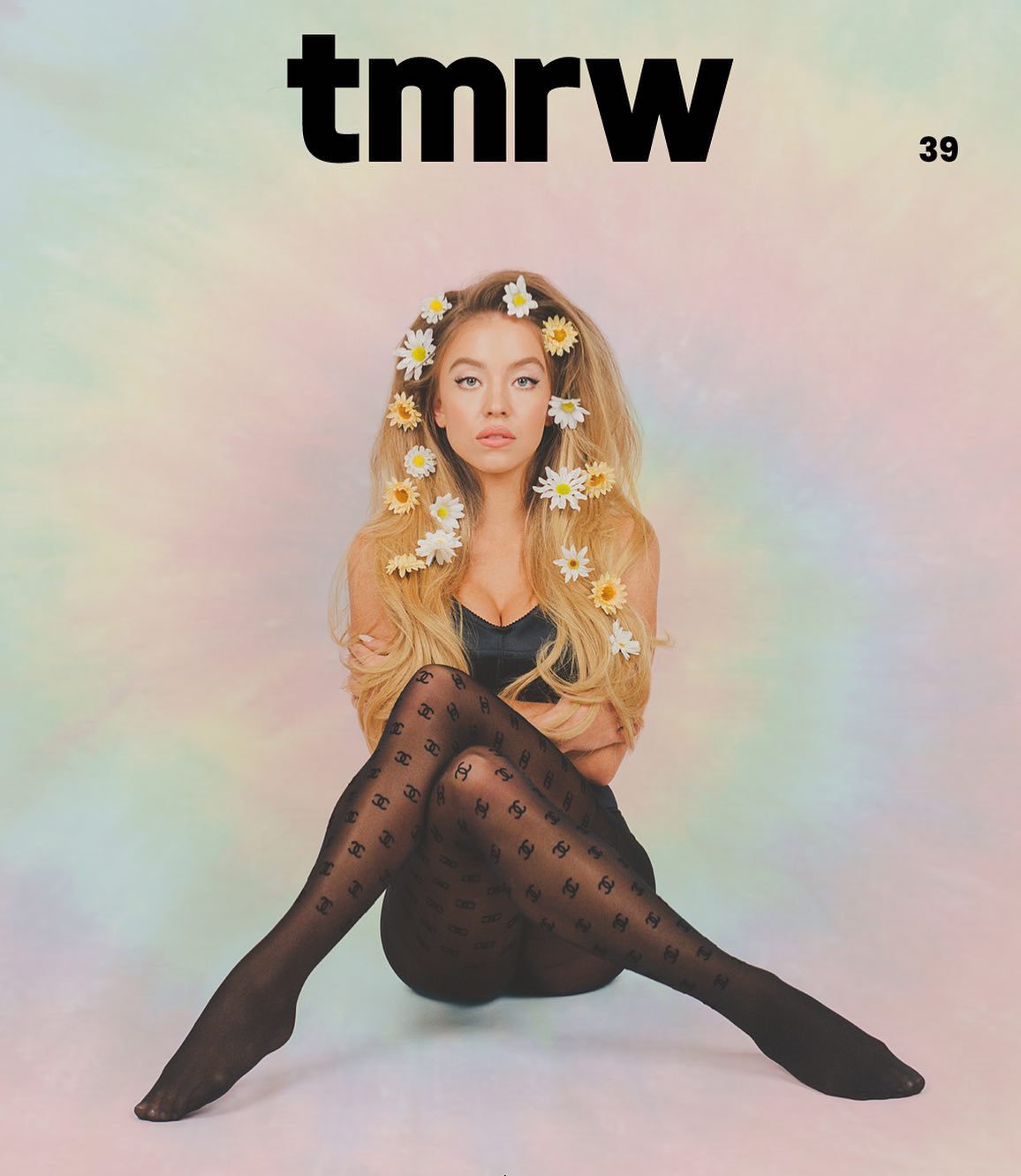 a cover shot from tmrw magazine, issue 39, featuring a young blonde woman with long hair adorned with daisy flowers. she is sitting with her legs crossed, wearing black sheer tights with a repeating chanel logo pattern. the background is a soft pastel color gradient. the overall style is fashionable and trendy.