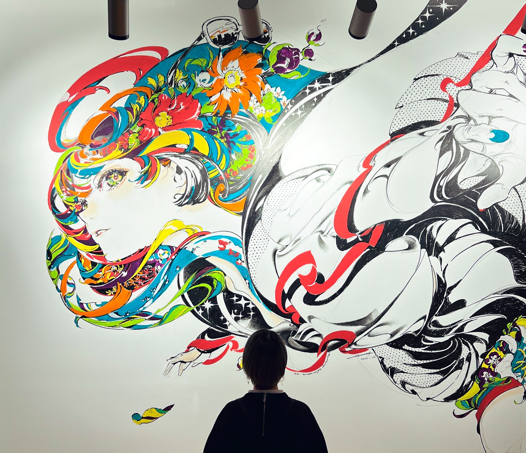 a woman stands before a large, colorful mural featuring abstract figures and floral patterns. the mural is brightly lit, highlighting the intricate details and vibrant colors. the woman's back is to the camera, emphasizing the scale and impact of the artwork. the style of the mural appears to be contemporary, combining traditional floral motifs with modern graphic elements.