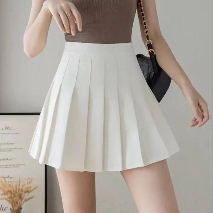 a young woman is standing in front of a white wall, wearing a white pleated mini skirt and a brown top. she is also wearing a black purse, and she is accessorizing with a ring on her finger. she is smiling and looking at the camera.