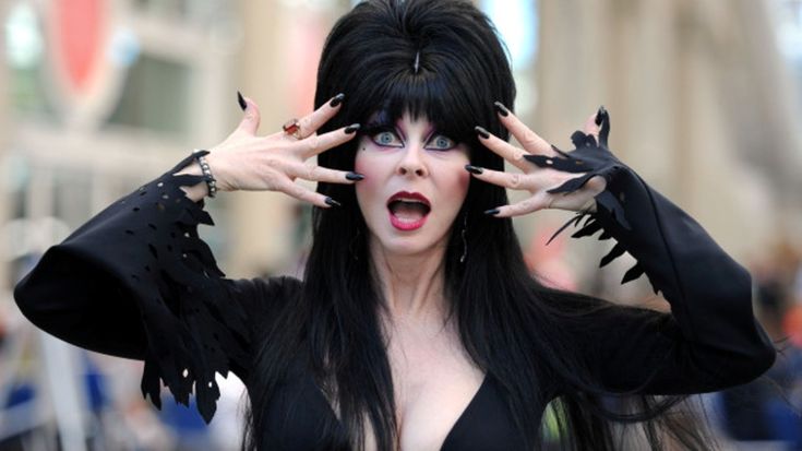 a woman with long black hair wearing a black dress and black makeup is smiling while looking at the camera. her nails are long and painted black. the image is cropped at the shoulders, showing her from the chest up.