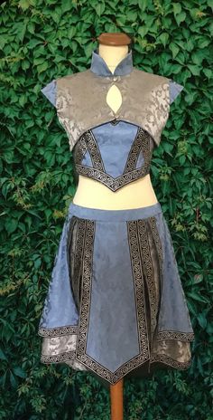 a blue and silver brocade warrior princess dress with gold trim is displayed on a mannequin. the dress is sleeveless and has a high collar. the skirt is short and flared. the mannequin is standing in front of a green hedge.