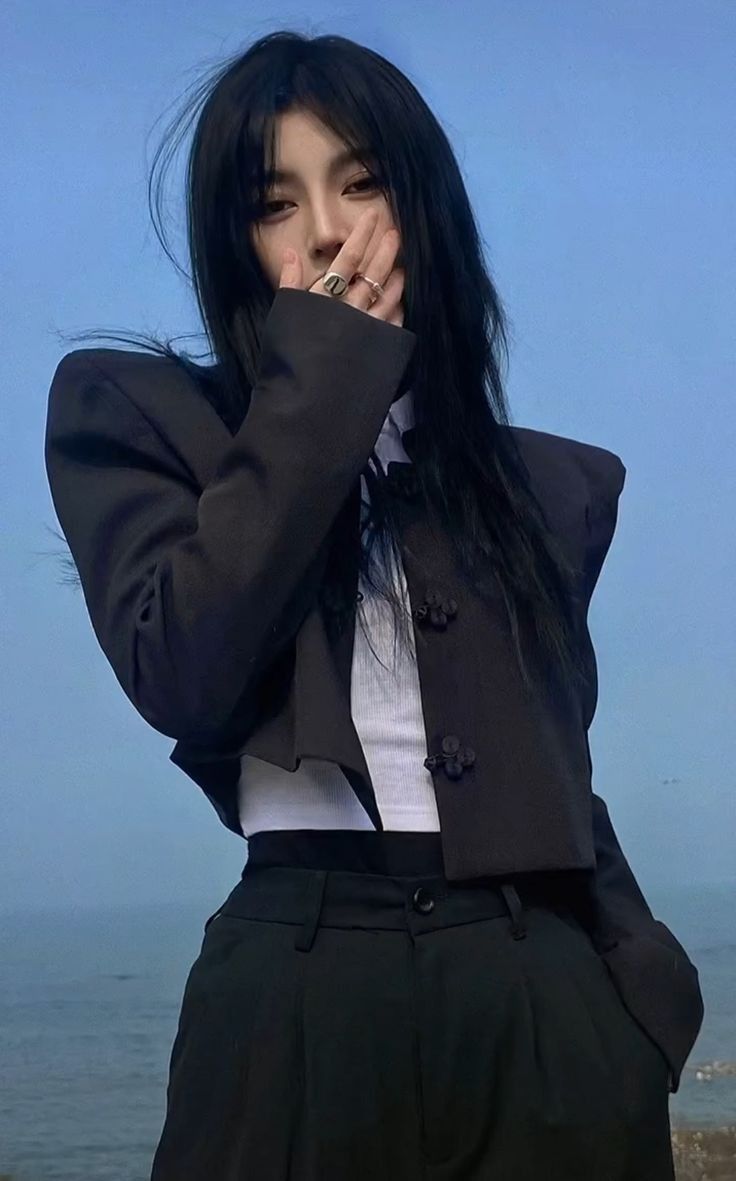 a young woman wearing a stylish black jacket and matching trousers stands outdoors with the ocean in the background. she has her hand over her mouth,  looking fashionable and cool.