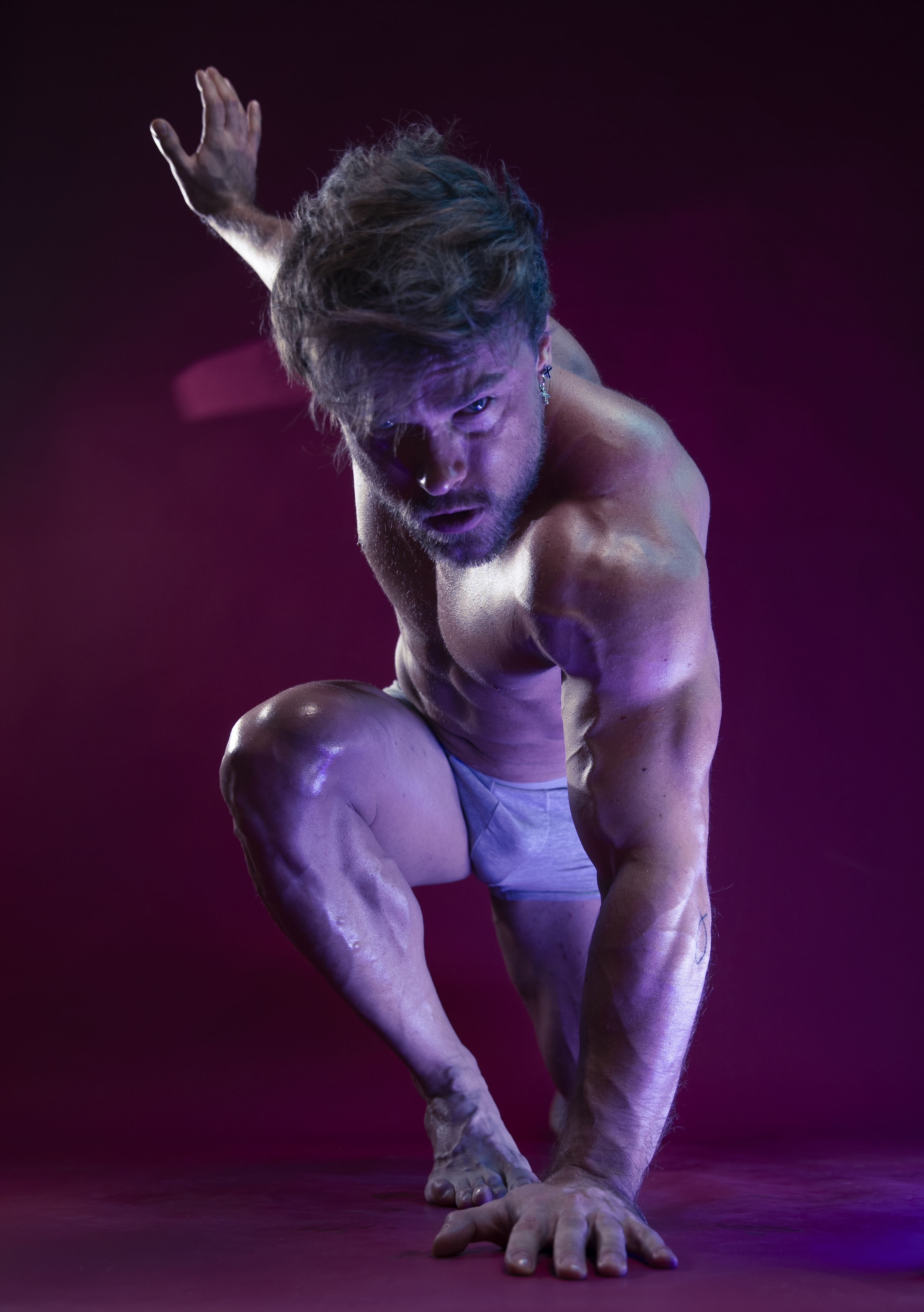 a muscular man is in a crouching pose with one hand on the ground and the other raised in the air. he is wearing underwear and is barefoot. the lighting is purple and the background is a dark red/purple color. his skin appears to be oiled, emphasizing his muscles. he is looking intensely at the viewer.