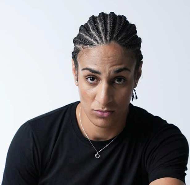 a woman with her hair in braids and wearing a black tshirt stares directly at the camera with a serious expression on her face. the image is a portrait taken indoors with soft lighting.