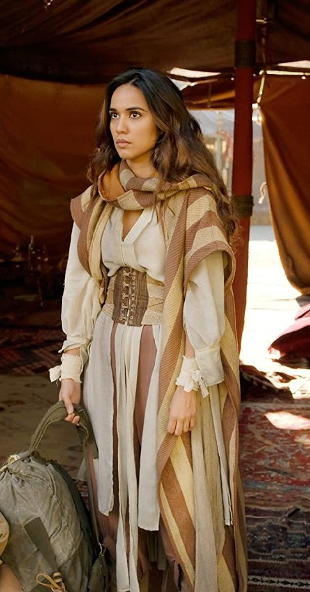 a woman in a long, beige and brown striped robe stands in a desert camp. she wears a corsetlike belt under her robe and has a scarf around her neck. she is carrying a large backpack. the setting appears to be ancient or historical. the woman looks thoughtful, gazing off to the side.