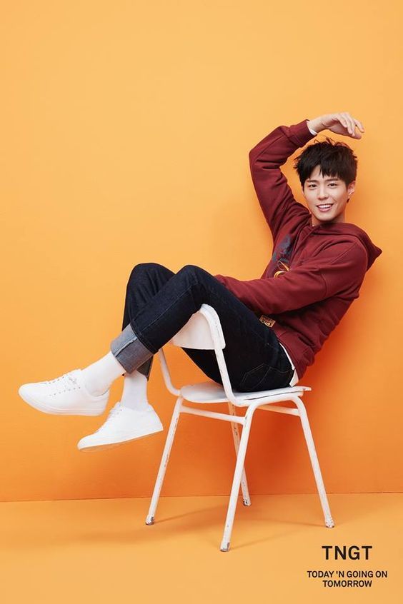 a man wearing a red hoodie and jeans is sitting on a white chair with his legs crossed. he is wearing white sneakers and is smiling at the camera. he is posing for a fashion photoshoot in front of an orange background.