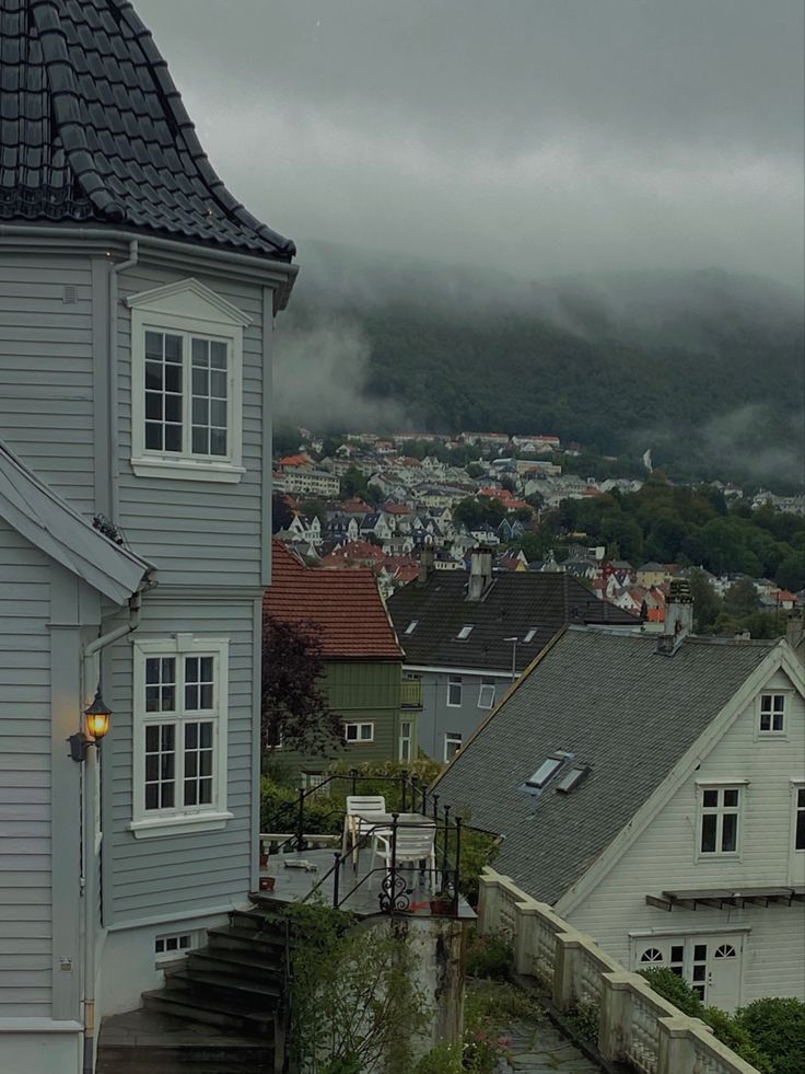 the image shows a grey house on a hillside overlooking a town nestled in misty mountains. the sky is overcast and it appears to be a rainy day. the architectural style of the houses is typical of northern europe. the main focus is on the grey house in the foreground, with its stairs leading up to a balcony and patio area. the background features many other houses cascading down the hill, obscured somewhat by fog. the overall atmosphere is calm and somewhat moody due to the weather and color palette.