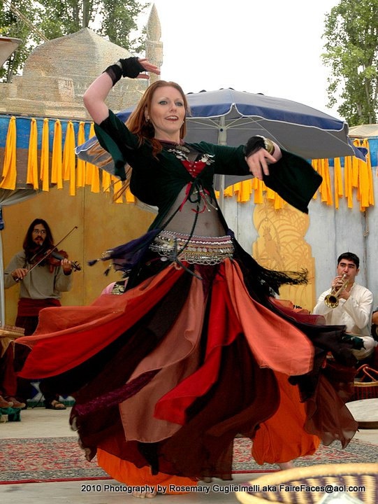 a full shot captures a woman in the midst of a belly dancing performance, set against the backdrop of an outdoor festival. the dancer, a fairskinned woman with reddish hair, is dressed in an elaborate costume. she wears a dark green top with flowing sleeves, a red bodice underneath, and a decorative headdress. her skirt is a vibrant display of multiple layers in shades of red, orange, and brown, creating a dynamic swirling effect as she moves. her arms are raised in a graceful dance pose, with black wristbands on both wrists and gold bracelets on one arm. behind the dancer, two men are playing musical instruments. to the left, a man with long dark hair and a beard plays the violin. to the right, another man in a lightcolored shirt plays the trumpet. they are both part of the performance, providing the music for the dancer's routine. the background includes elements of a festival setting, such as colorful banners, tents with blue and white canopies, and a carpet on the ground. in the distance, there is a domelike structure, suggesting an exotic or middle eastern theme. the overall atmosphere is lively and festive, with the focus on the dancer and her mesmerizing performance.