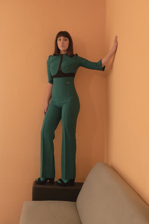 a woman is standing on top of a black block, which is on a gray sofa. she is wearing a green jumpsuit with black detailing around the chest. she is posing with one hand against the orange wall behind her.