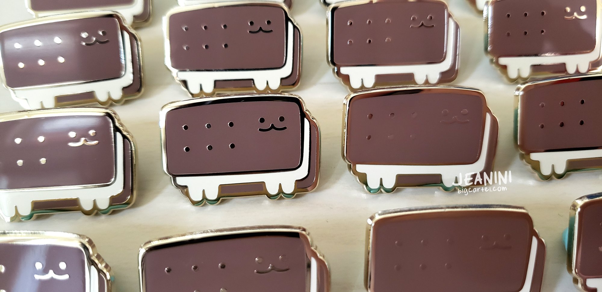 a closeup shot of multiple ice cream sandwich enamel pins laid out on a white surface. the pins feature a cute cartoonlike cat design on a chocolate ice cream sandwich. the pins are brown, white, and gold colored with a shiny finish.  the pins are all similar in design, but have slight variations in detail.  the background is a blurred out white surface. a watermark is visible in the lower right corner with text “jeanini bigcartel.com”.