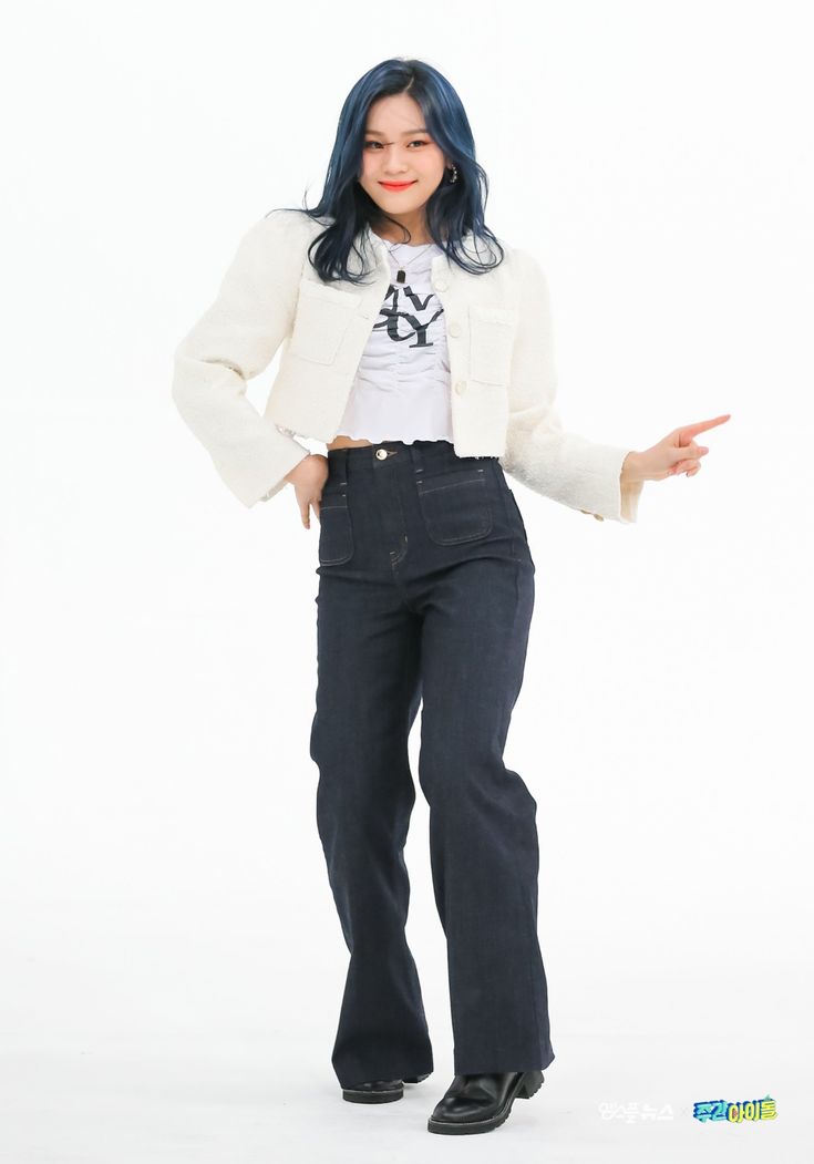 a young woman with blue hair is posing in a white cropped jacket, dark blue flare jeans, and black boots. she is smiling and looking at the camera. the background is a white studio wall.