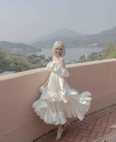 a young woman with long blonde hair is wearing a white frilly dress and white heels. she is standing in front of a wall overlooking a scenic mountain landscape with a lake in the distance. she is smiling and looks happy. the image is likely taken during the summer.