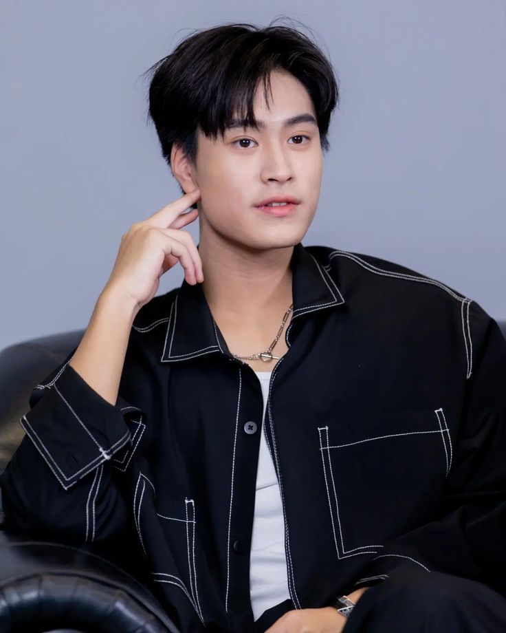 a young asian man is sitting on a black leather couch. he is wearing a black jacket with white stitching and a white shirt. he has short black hair and is looking at the camera. the background is a light gray wall. the lighting is soft and the image is taken from eye level.