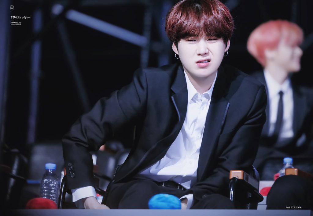 a man in a dark suit with a white shirt is sitting and smiling at the camera. he is surrounded by other people. the background is blurred. there are water bottles on the table in front of him. he is holding a microphone. he is wearing a red hair and has an earing.