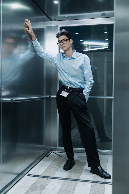a stylish young man in a light blue shirt and black pants stands in an elevator, leaning against the wall. he's wearing glasses and has a confident pose. the elevator's metal interior is reflected in the shiny walls. the overall atmosphere is modern and professional.