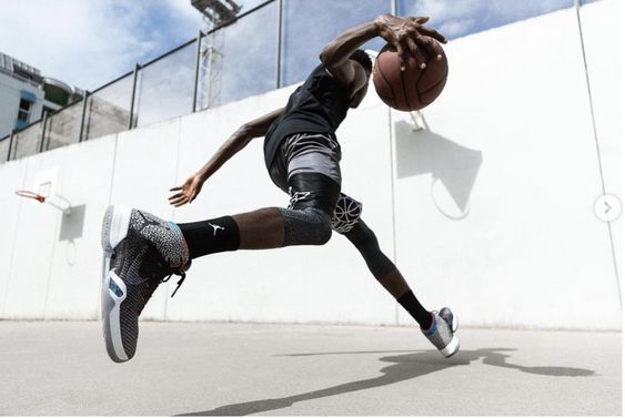 a basketball player in midair, executing a jump shot on an outdoor court. he's wearing athletic apparel, including basketball shoes, and his pose captures the energy and dynamism of the sport. the bright, sunny day illuminates the scene, while a low angle shot emphasizes the player's height and athleticism.