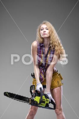 a beautiful blonde woman holds a chainsaw in front of her. she is wearing a plaid shirt, shorts, and suspenders. she is looking at the camera with a serious expression on her face.