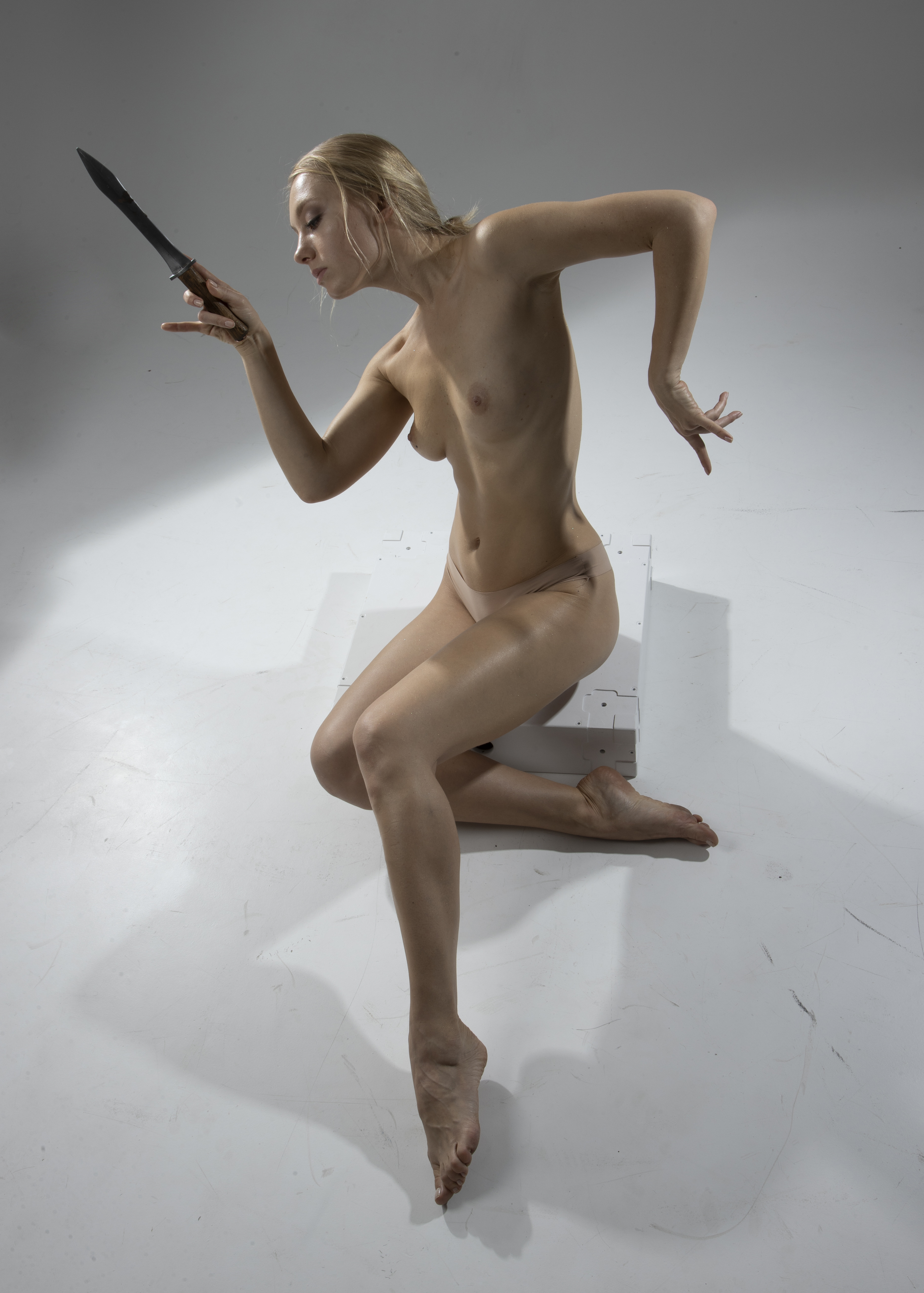 a nude fairskinned woman with blonde hair is sitting on a white surface, holding a knife in her right hand. she is looking at the knife. the background is a plain white. the woman's body is illuminated by a soft light. her left hand is extended out to her side, fingers splayed. she is sitting with her legs crossed.